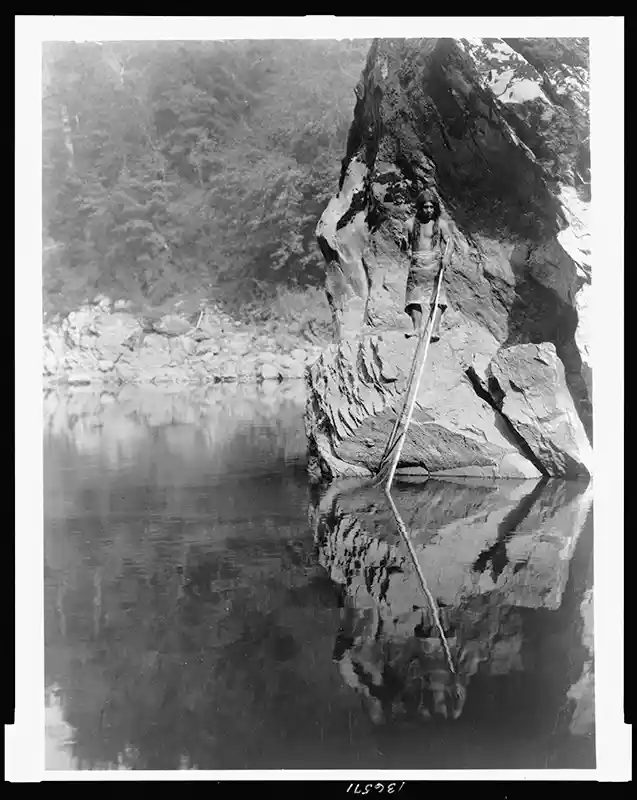 Yurok man standing on rock ledge holding fishing line in one hand and pole down into the water, c1923, Curtis, Edward S., 1868-1952